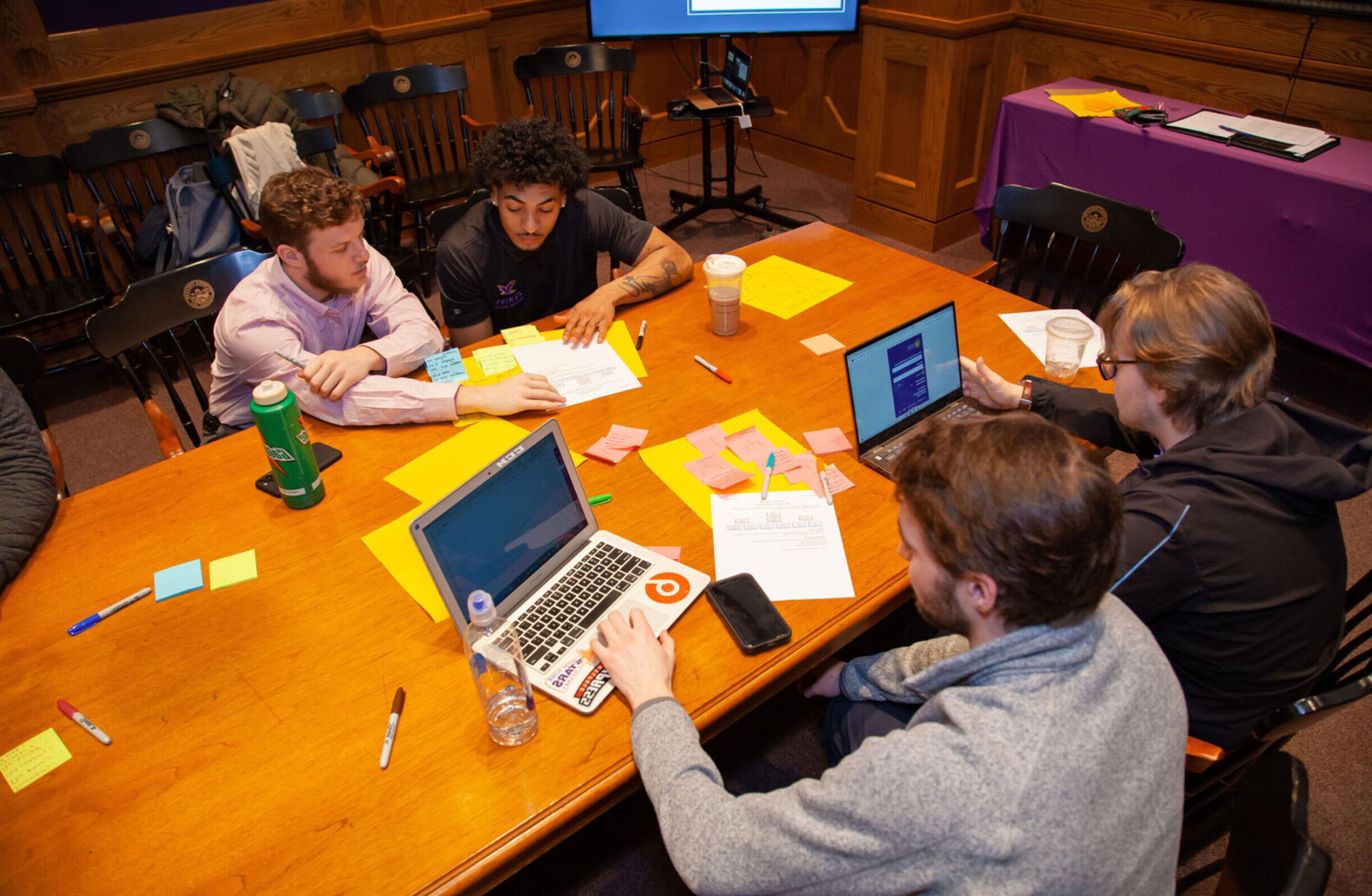 Two male students work on laptops while two other male students work together around a wooden table